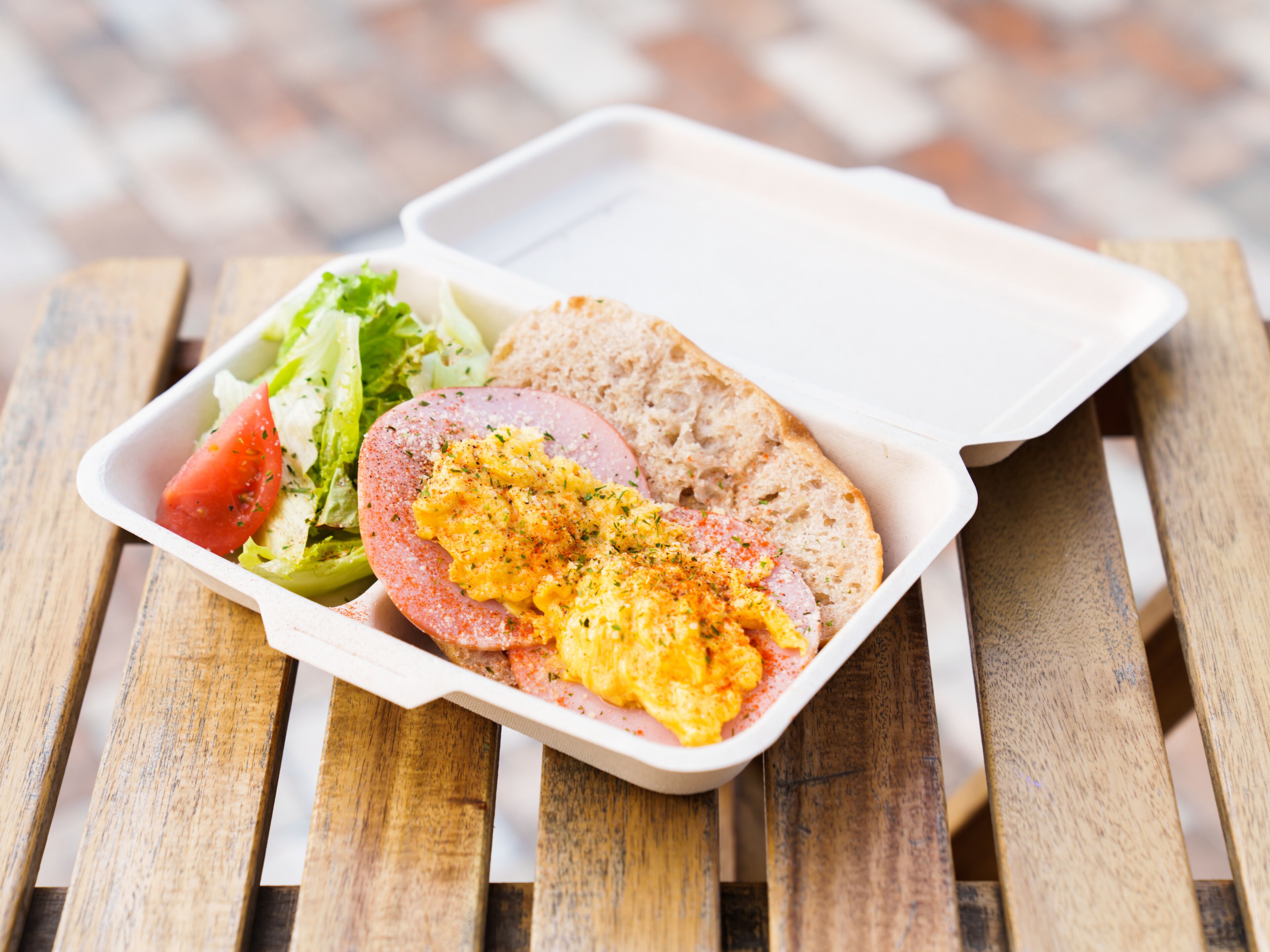 Eine Mahlzeit in einem weißen Einwegbehälter bestehend aus Rührei, einer Scheibe Schinken, einer Scheibe Brot und einem Beilagensalat auf einem Holztisch.