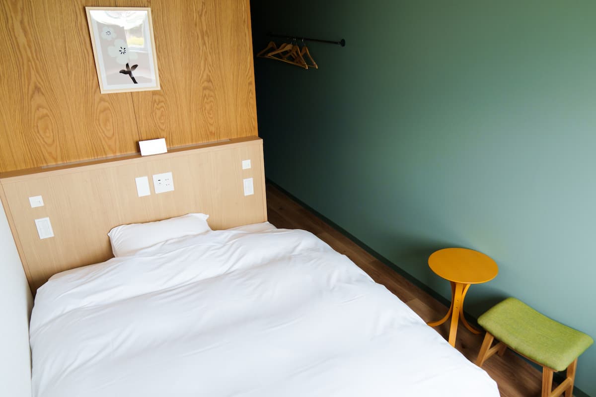 Un lit bien fait dans une pièce dotée d’une tête de lit en bois, d’une petite table ronde avec un banc vert et d’un porte-manteau fixé au mur.