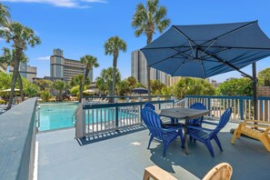 Un área de piscina en la azotea con una mesa y sillas, rodeada de palmeras y un horizonte urbano.