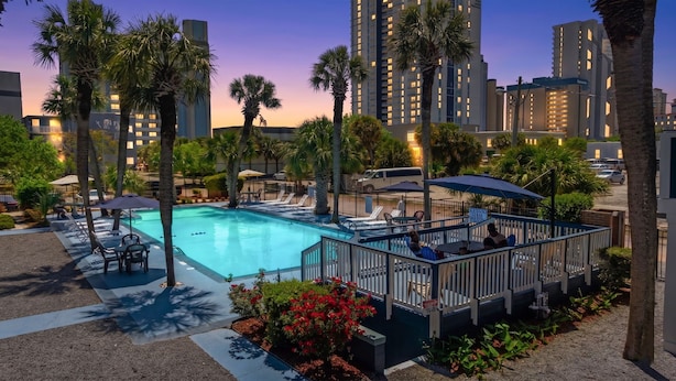 A swimming pool with lounge chairs and umbrellas, surrounded by palm trees and a city skyline in the background. - Sun Fun Motel (Myrtle Beach)
