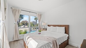 A bedroom with a large window, a bed with white bedding, a bedside table with a lamp, and a view of the outside. - Sun Fun Motel (Myrtle Beach)
