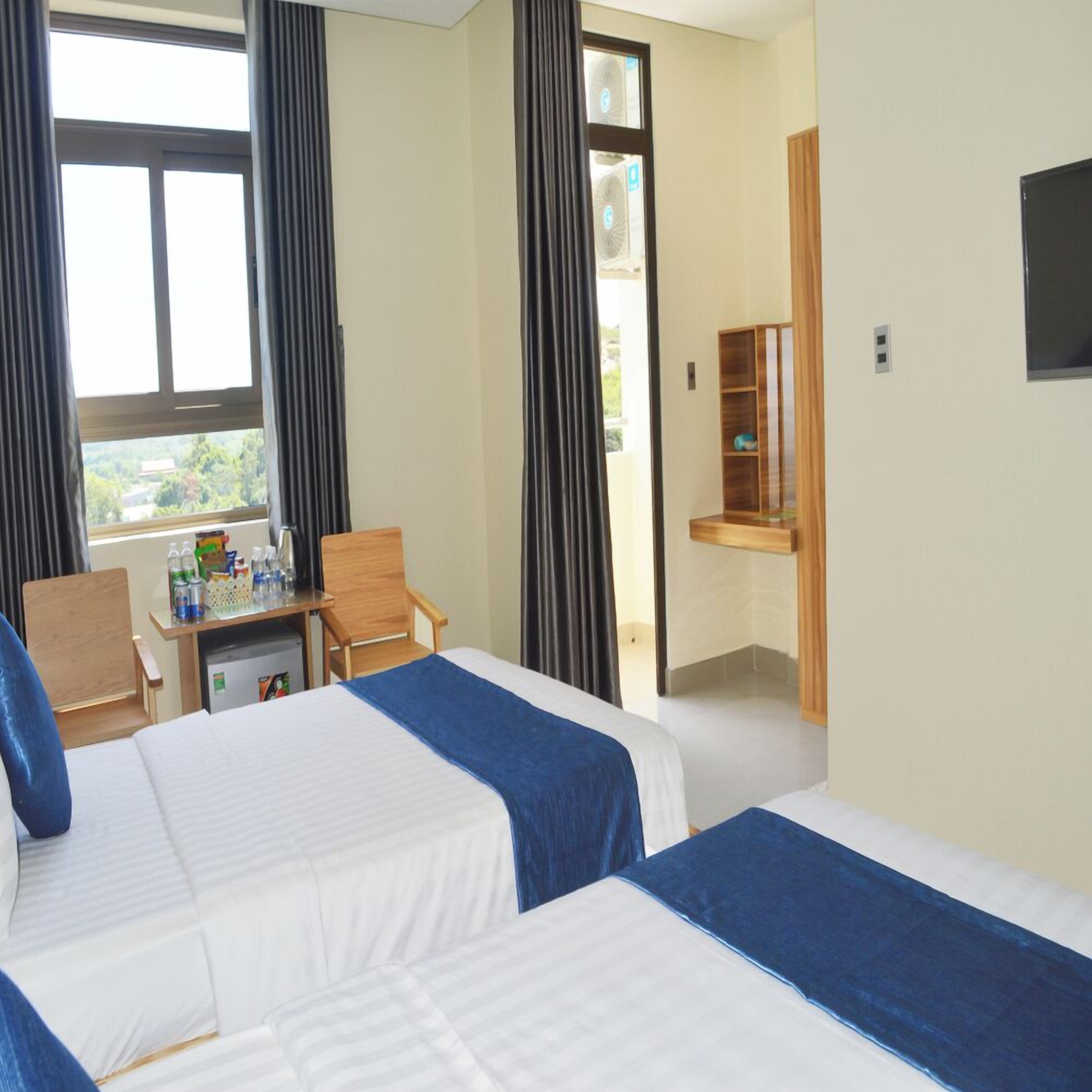 A hotel room with two beds, a desk with a chair, a small kitchenette, and a window with curtains.