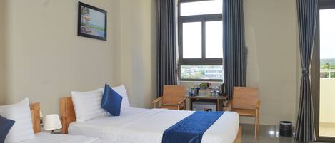 A hotel room with two beds, a desk, chairs, a window with curtains, and a small table with items on it.