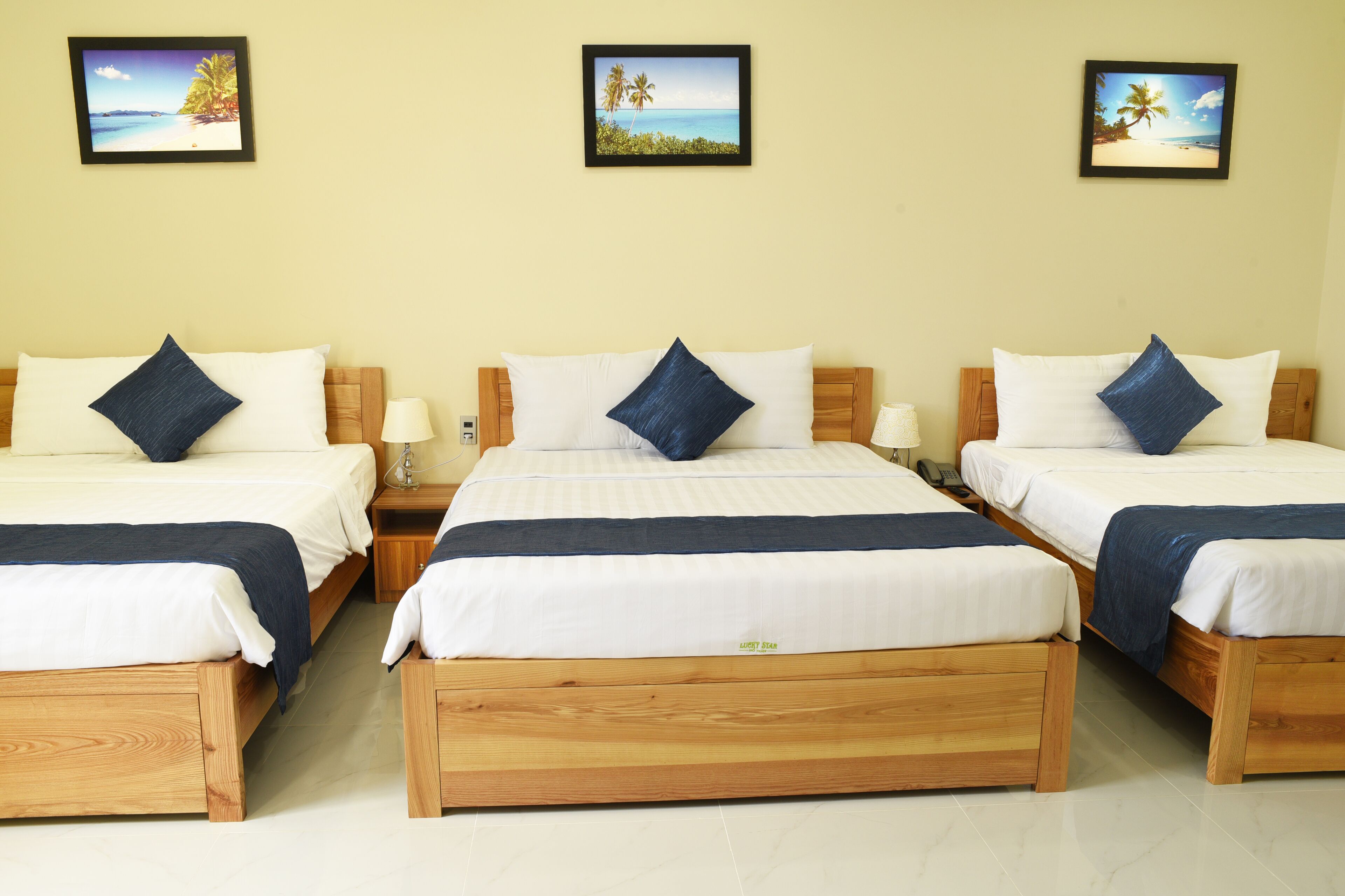A hotel room with three wooden beds, each with white bedding and blue pillows, and framed tropical beach pictures on the wall.