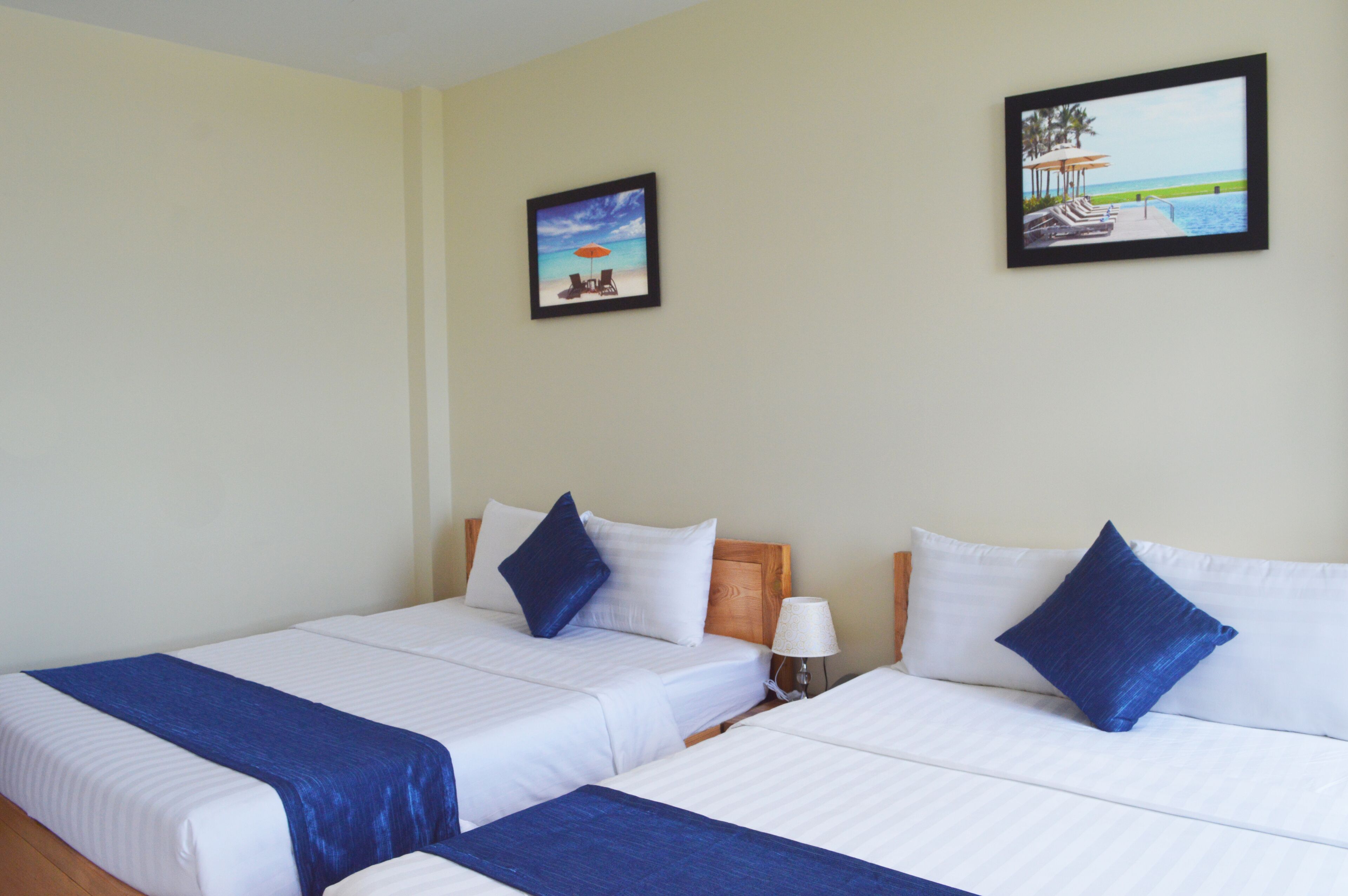 Una habitación de hotel con dos camas, cada una con una almohada azul decorativa, una mesita de noche con lámpara, y dos cuadros enmarcados en la pared.