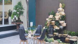 Un espace patio aménagé avec une table en verre et quatre chaises, entouré d’une cascade artificielle en roche et de divers plantes en pot.