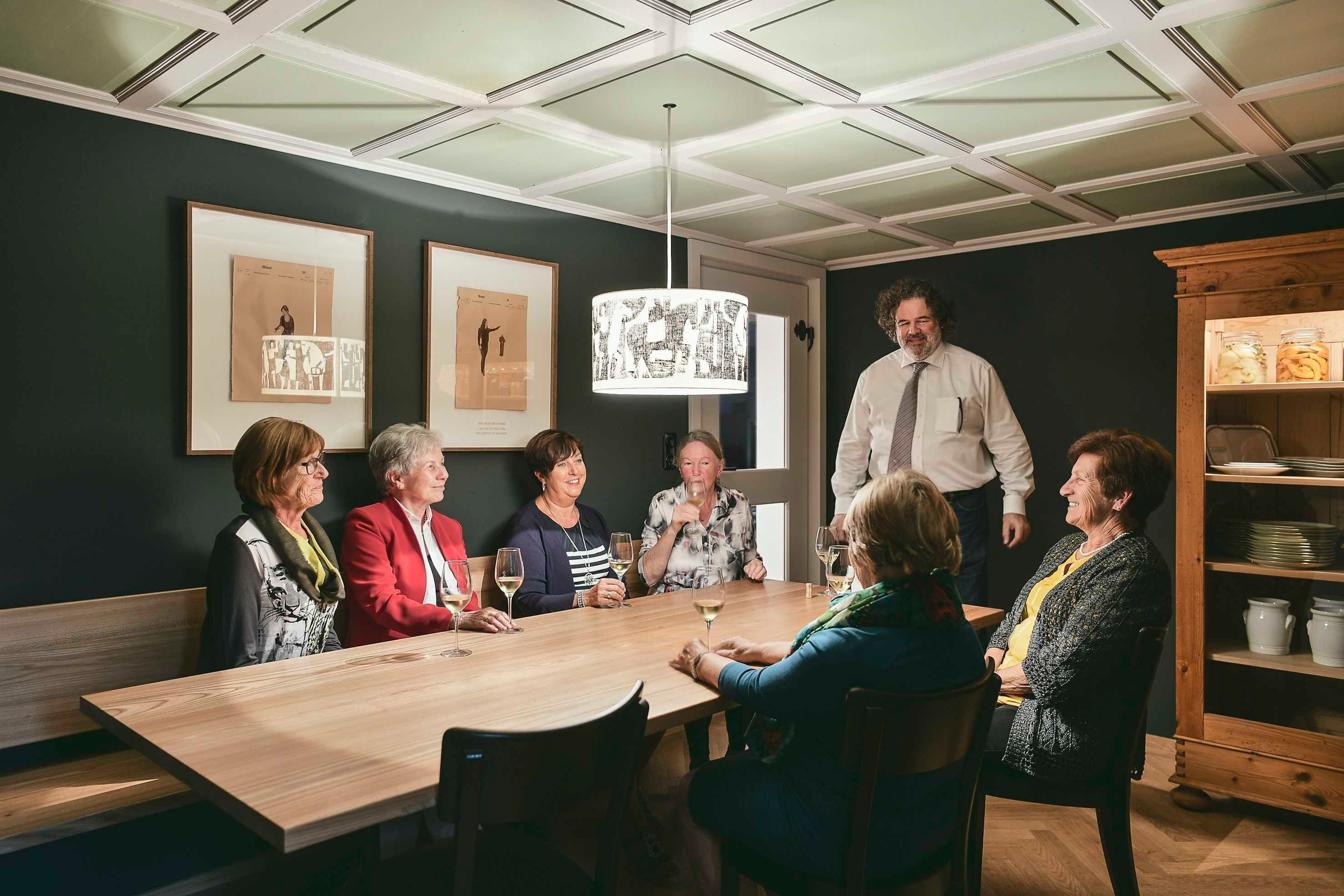 Un groupe de personnes est assis autour d’une table en bois dans une pièce au plafond haut, dont les murs sont ornés d’œuvres d’art encadrées.