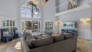 A spacious living room with a large gray sofa, two armchairs, a coffee table, and a TV on a stand. There are large windows with shutters, a ceiling fan, and a staircase to an upper level. - DIRECT Gulf access, Heated Pool & Spa, Kayaks - Dance the night away at Villa Dancing Flamingo! (Cape Coral)