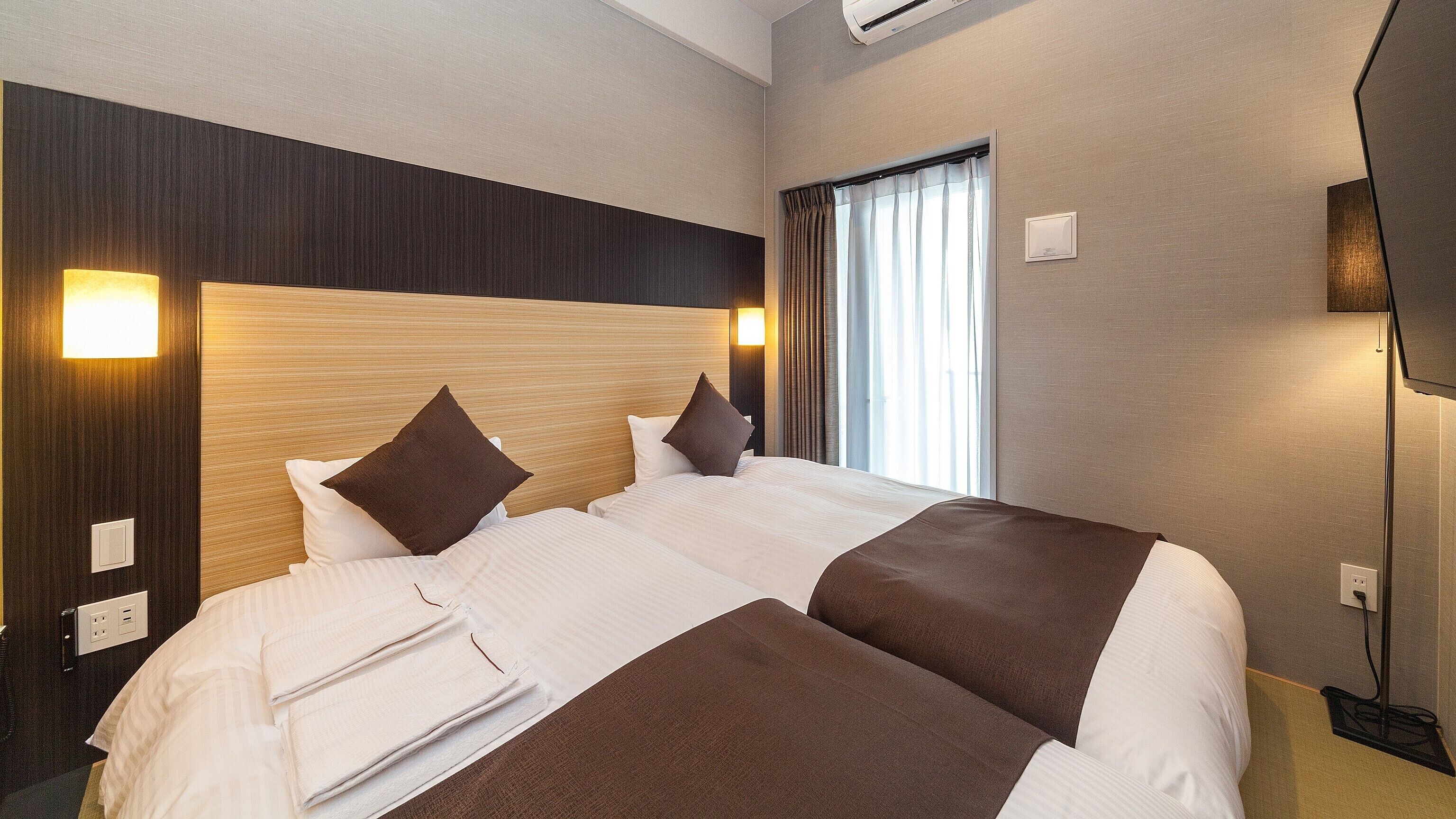 Ein Hotelzimmer mit einem großen Bett, einem Kopfteil, einem Fenster mit Vorhängen, einem an der Wand montierten Fernseher und einem Nachttisch mit einer Lampe.