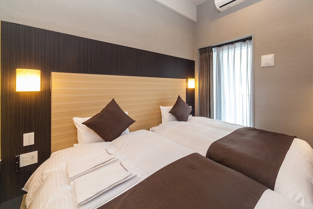 Ein Hotelzimmer mit einem großen Bett, einem Kopfteil, einem Fenster mit Vorhängen, einem an der Wand montierten Fernseher und einem Nachttisch mit einer Lampe.