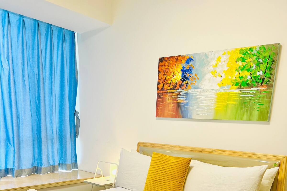 Een net opgemaakt bed met witte en gele lakens, een houten hoofdbord en een schilderij van bomen aan de muur.