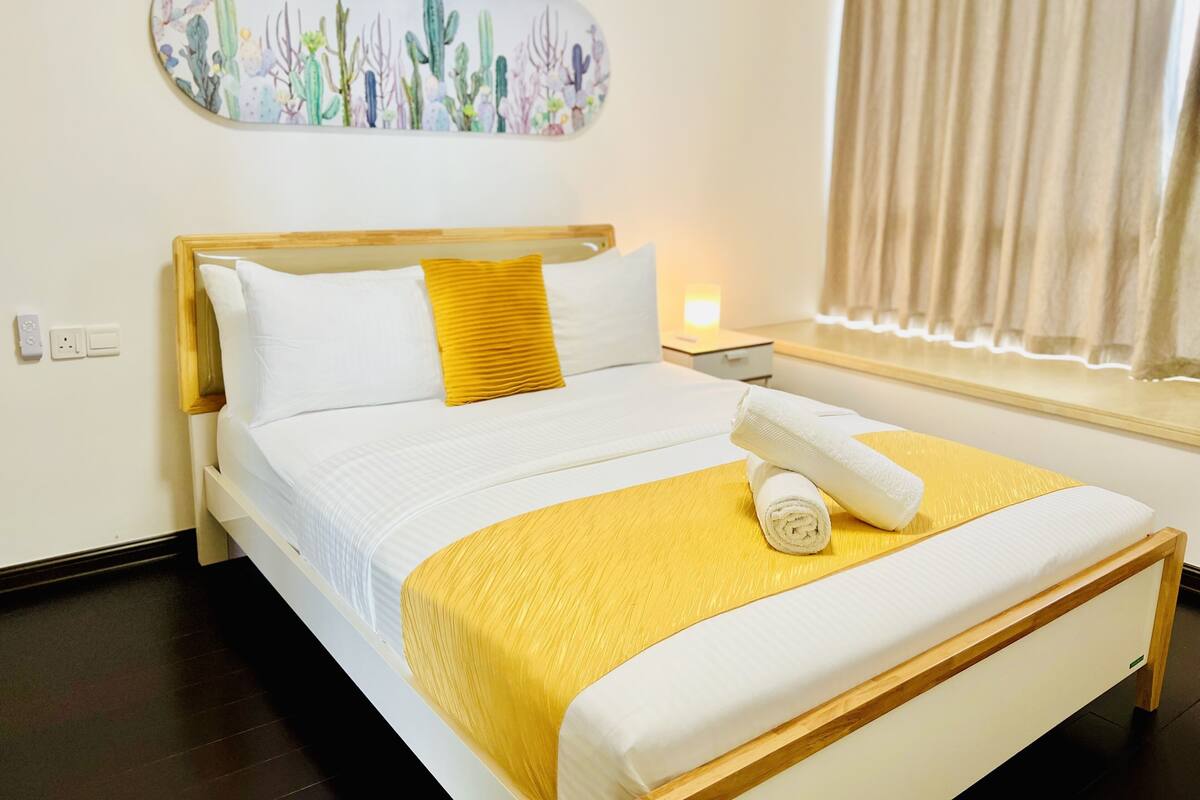 Een net opgemaakt bed met wit en geel beddengoed, een houten hoofdbord en een wanddecoratie van cactussen.