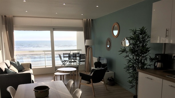 Ein modernes Wohnzimmer mit großem Fenster mit Meerblick, einem Essbereich mit Tisch und Stühlen, einem Sofa, einem kleinen runden Tisch und einer Küchenzeile mit Schränken und einer Kaffeemaschine.
