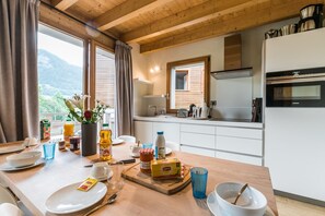 Una mesa de comedor de madera dispuesta con platos, cubiertos y condimentos, una planta en maceta y vistas de montañas a través de amplios ventanales.