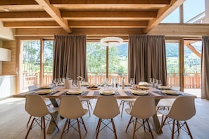 Uma sala de jantar com uma mesa de madeira preparada para seis pessoas, grandes janelas e vista para o exterior.