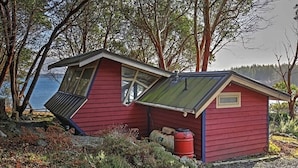 A small, red wooden cabin with a sloped roof, a window, and a door, situated by a body of water with trees in the background. - Studio with a View!! (Friday Harbor)