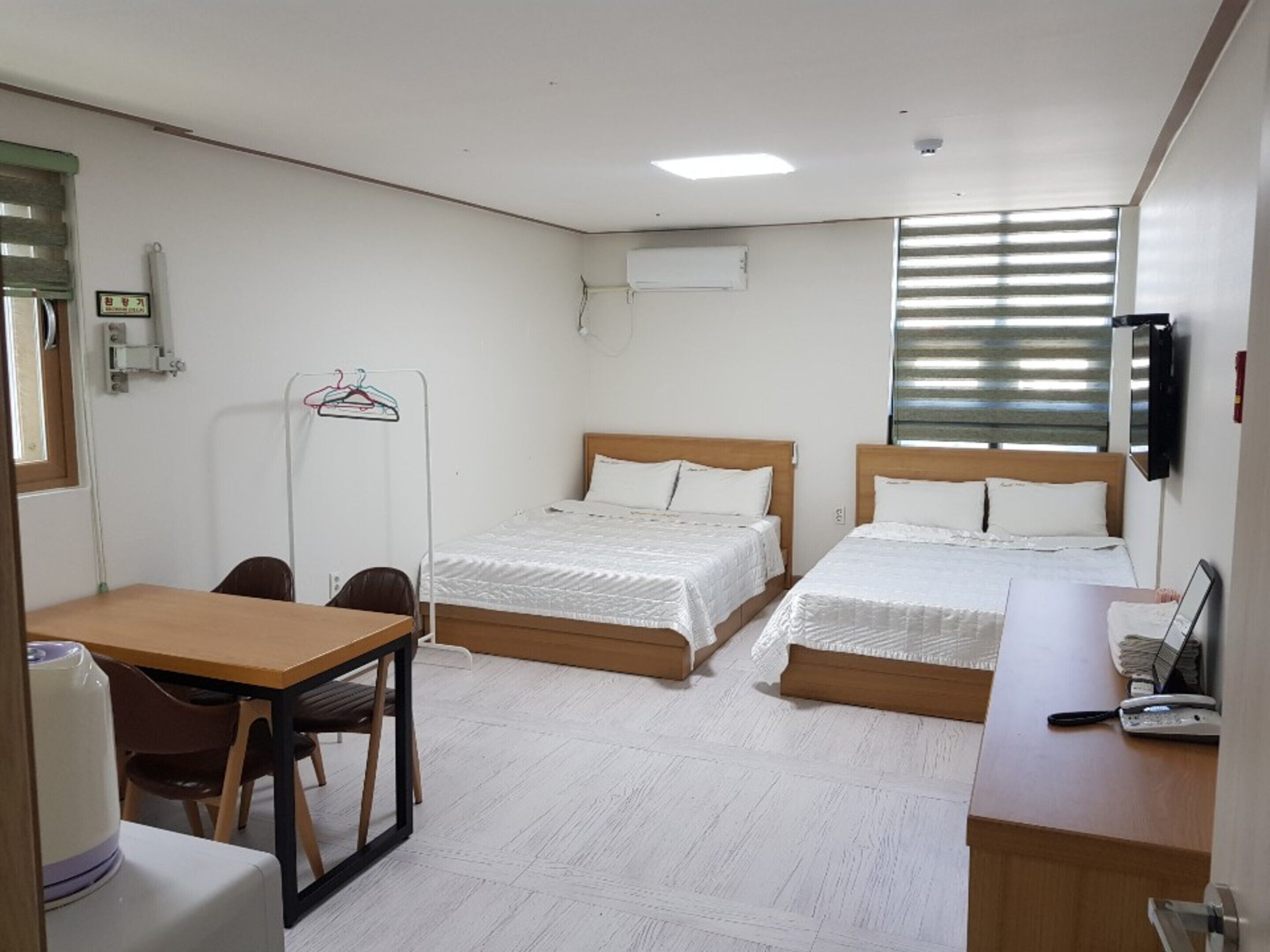 A hotel room with two beds, a desk, and a chair. There is a window with blinds, a TV mounted on the wall, and a small table with a kettle.