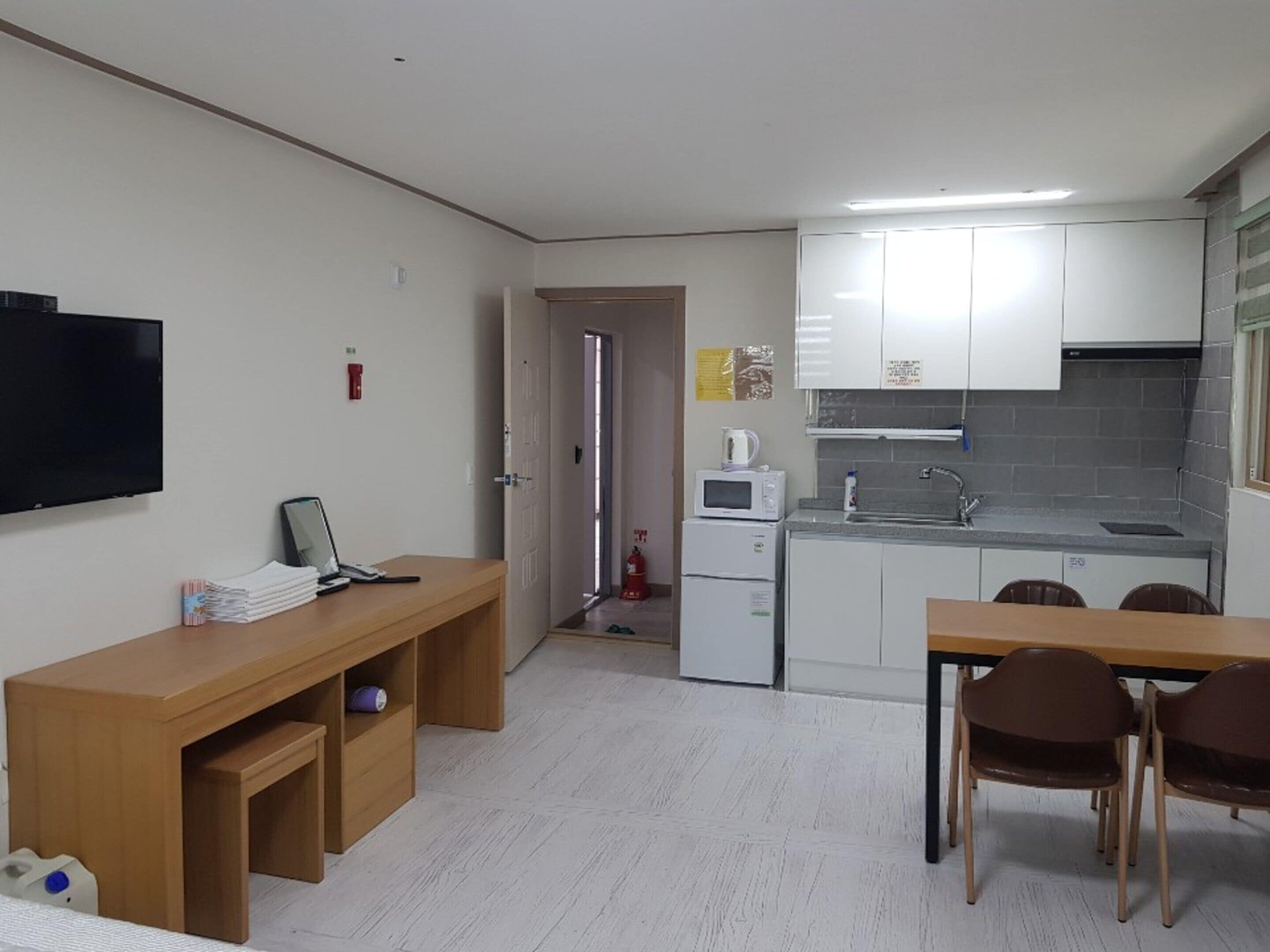 Cocina compacta con armarios blancos, microondas y fregadero. Hay una mesa y sillas de madera en la zona de comedor, y un televisor montado en la pared.