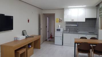 Cocina compacta con armarios blancos, microondas y fregadero. Hay una mesa y sillas de madera en la zona de comedor, y un televisor montado en la pared.