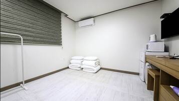 Et værelse med hvide fliser, en radiator, et foldet hvidt håndklæde, en mikroovn og et træskrivebord med et fjernsyn.