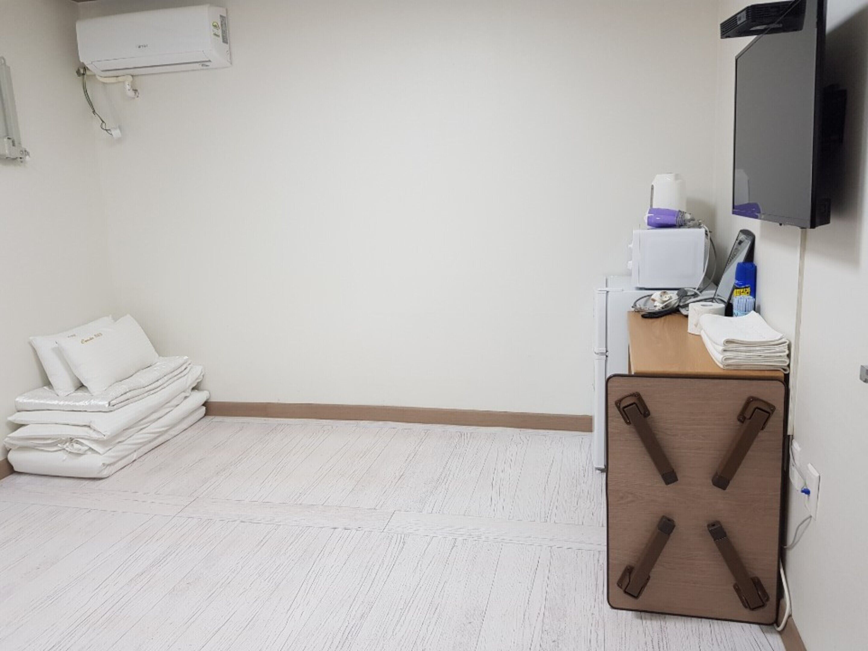 Une salle de consultation avec un sol blanc, une pile de linge blanc, un meuble en bois avec un plateau, et un téléviseur fixé au mur.