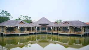 A series of thatched-roof buildings with wooden railings over a calm body of water, reflecting the structures. - Memorina Ninh Binh Resort (Tay Hoa Lu)