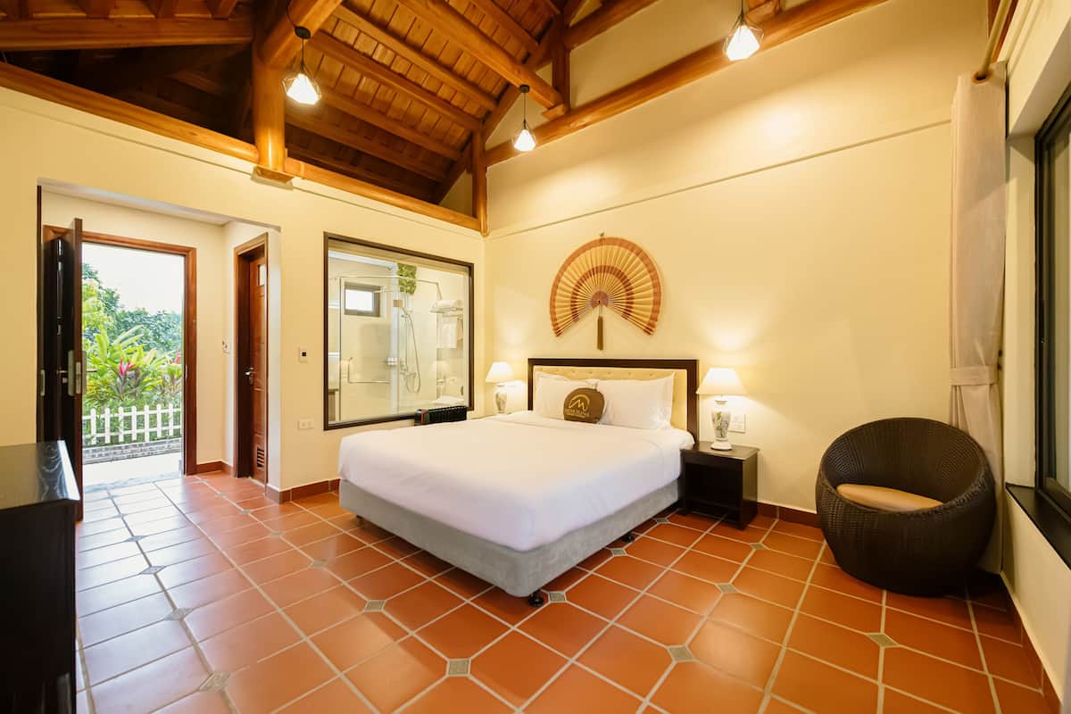 A bedroom with a large bed, a nightstand, a wicker chair, and a fan on the wall.