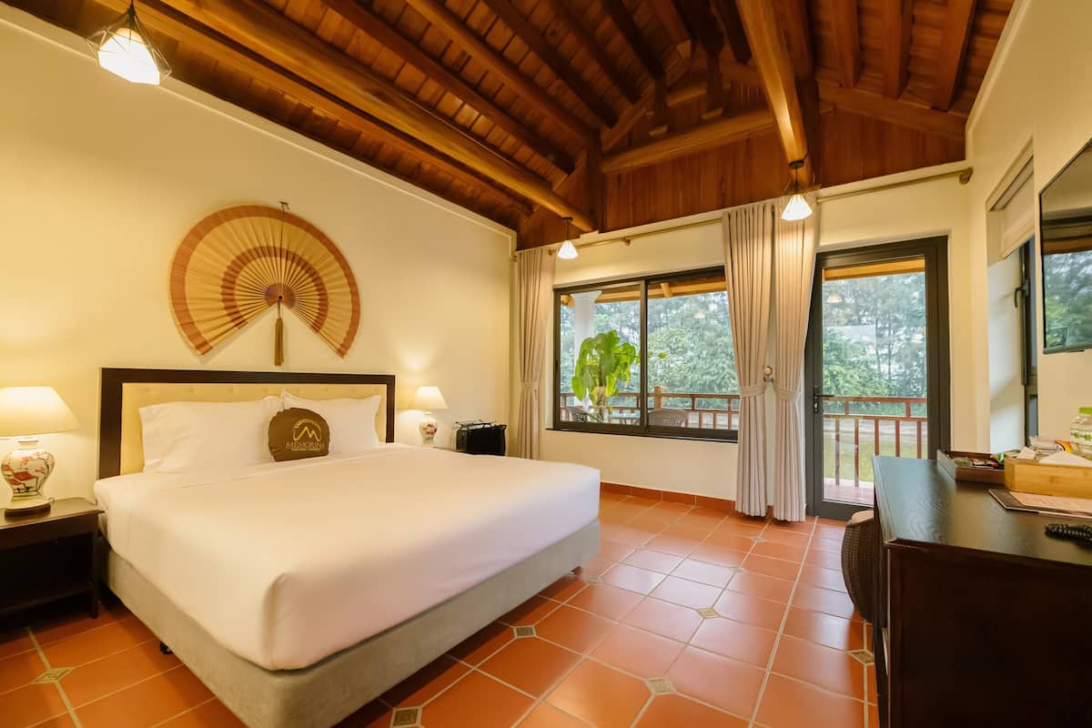 A hotel room with a large bed, wooden ceiling, a fan, a desk, and a window with curtains.