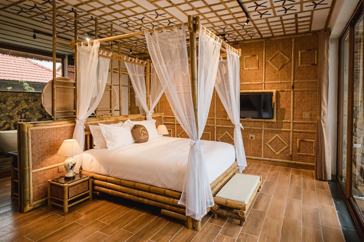 A four-poster bed with white bedding and a canopy, a bedside table with a lamp, and a stone wall with a TV.