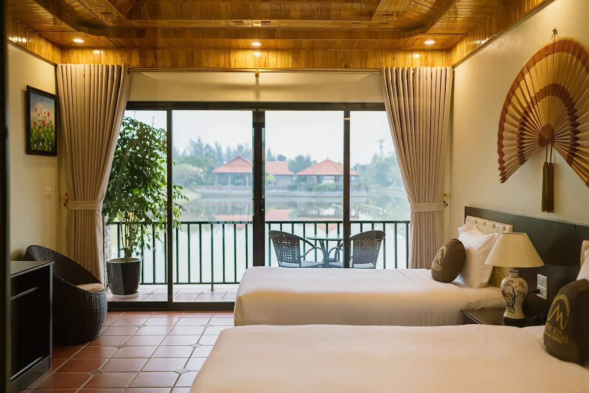 A hotel room with two beds, a balcony with a table and chairs, a potted plant, and a view of buildings.