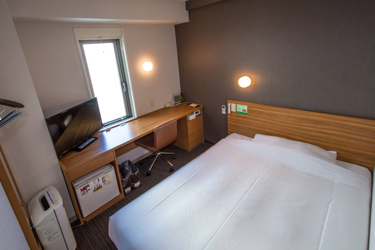 Hotelkamer met een bed, een bureau, een stoel, een kleine koelkast, een prullenbak en een raam.