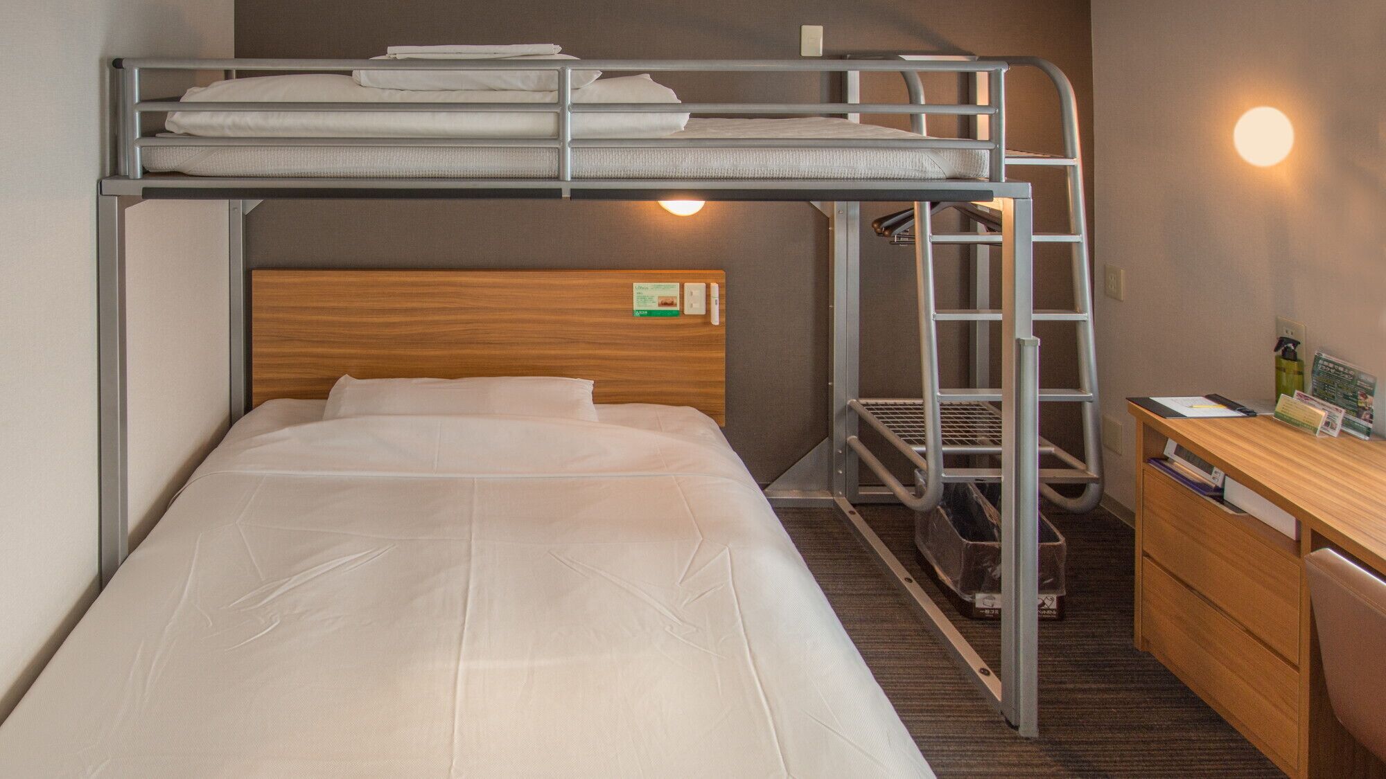 A bunk bed with a desk and a chair, a wooden headboard, and a wall-mounted light.