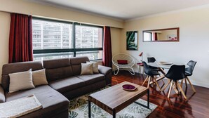 Un salon moderne avec un canapé marron, une table à manger avec des chaises, une grande fenêtre avec des rideaux et un luminaire au plafond.