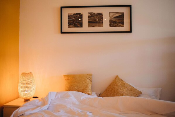 A neatly made bed with white linens and two patterned pillows, a framed photo of a bridge, and a lamp on a bedside table. - Budget-Friendly Studio w/ Balcony in Rosario (Porto)