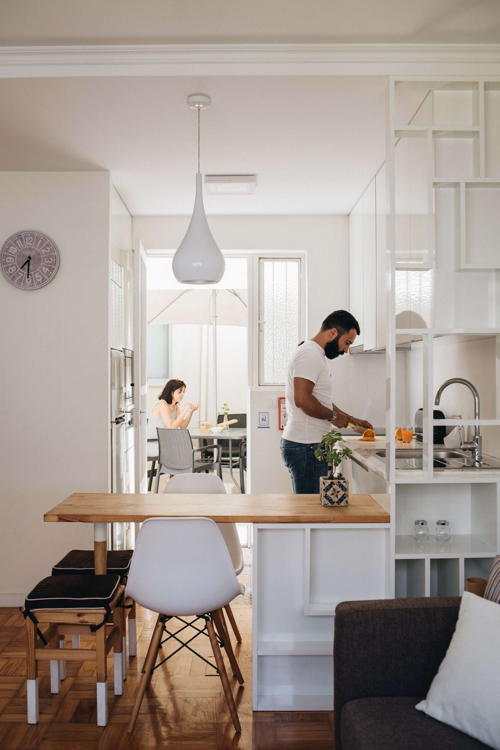 A modern kitchen with white cabinetry, a wooden dining table, and a person preparing food.
