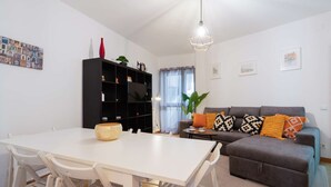 Uma sala de estar moderna com uma mesa de jantar branca, um sofá cinza com almofadas coloridas, uma estante preta e um lustre.