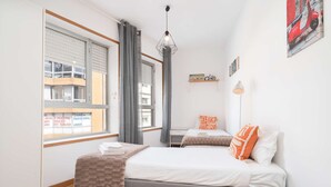 Quarto com duas camas, janela com vista para edifícios, luminária pendente e prateleira na parede com um modelo de bicicleta.