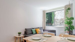 Een eetgedeelte met een tafel voor vier personen, een grijze bank, een klein bijzettafeltje met een plant en een raam met uitzicht op een stenen muur.