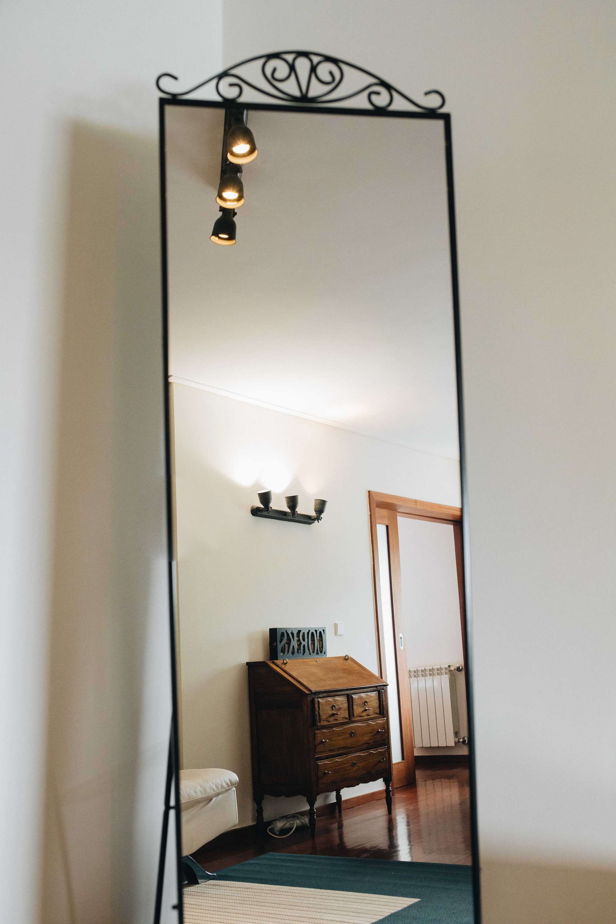 A tall, narrow mirror with a decorative top and three light fixtures. A wooden dresser with drawers is visible in the reflection.