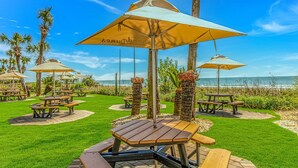A beachside patio with picnic tables and umbrellas, palm trees, and a view of the ocean.
