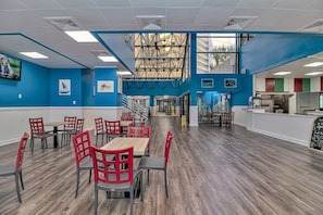 Un vaste espace dédié à la salle à manger, avec plusieurs tables et chaises, un escalier menant à un niveau supérieur et une vue sur l’extérieur grâce à de grandes fenêtres.