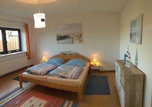 A bedroom with a bed, two bedside lamps, a wooden dresser, a window with curtains, and a wall-mounted painting.
