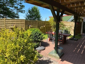 A covered patio area with a table and chairs, surrounded by greenery and a wooden fence.