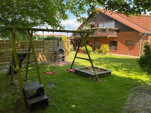 Un jardin avec une balançoire, une échelle en bois, un petit marchepied et une maison en briques avec un toit rouge.