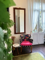 Una habitación con un espejo de marco de madera, una silla de lunares con un cojín floral, una pequeña mesa redonda y una alfombra verde estampada.