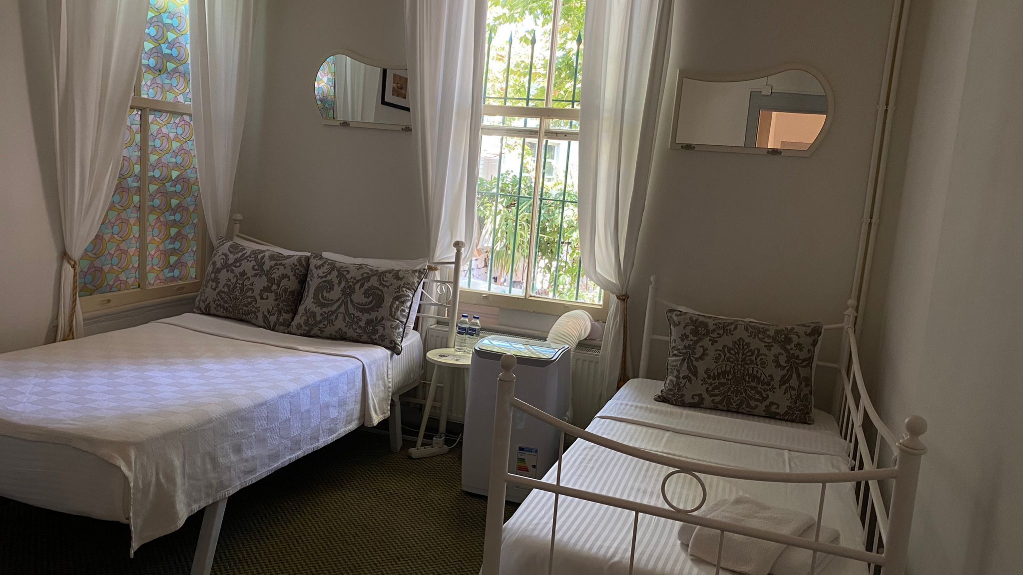 A bedroom with two beds, a small table, a chair, a radiator, and a window with a view of greenery.