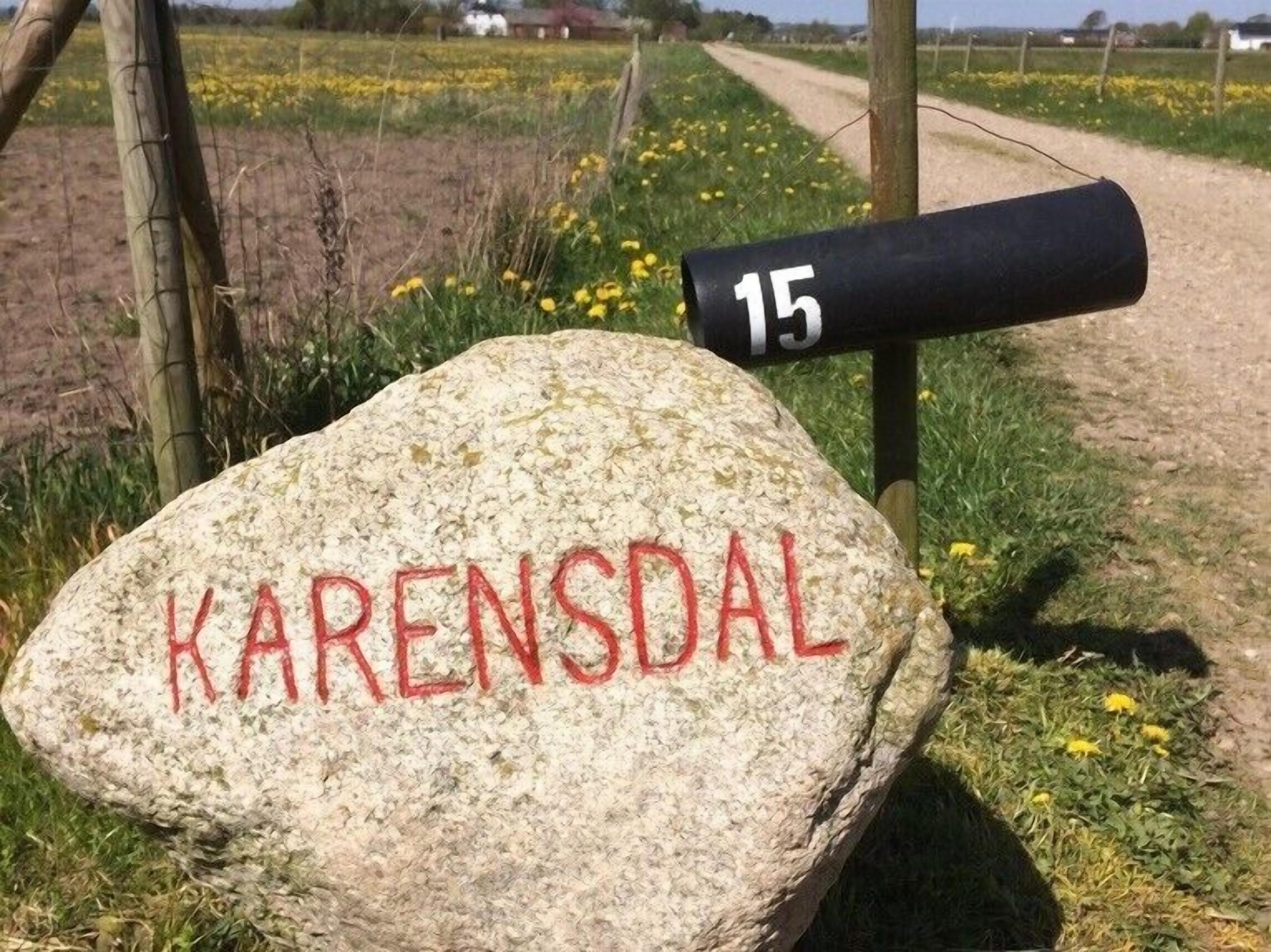 Une pierre sur laquelle est gravé le mot « KARENSDAL », un poteau noir portant le numéro « 15 », et un paysage rural avec des champs et un chemin de terre.