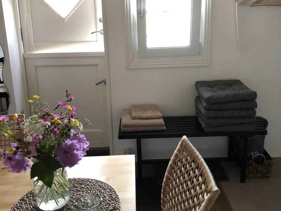 A room with a wooden table, a wicker chair, a window with a view, and a bench with folded towels.