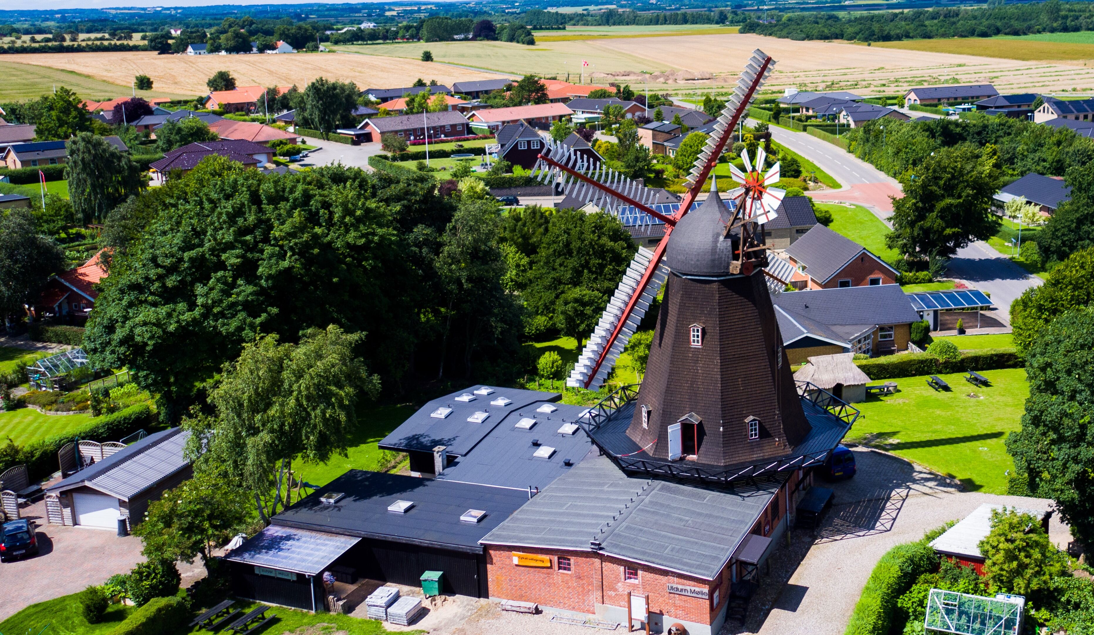 A windmill with a modern extension, surrounded by residential houses and green fields.