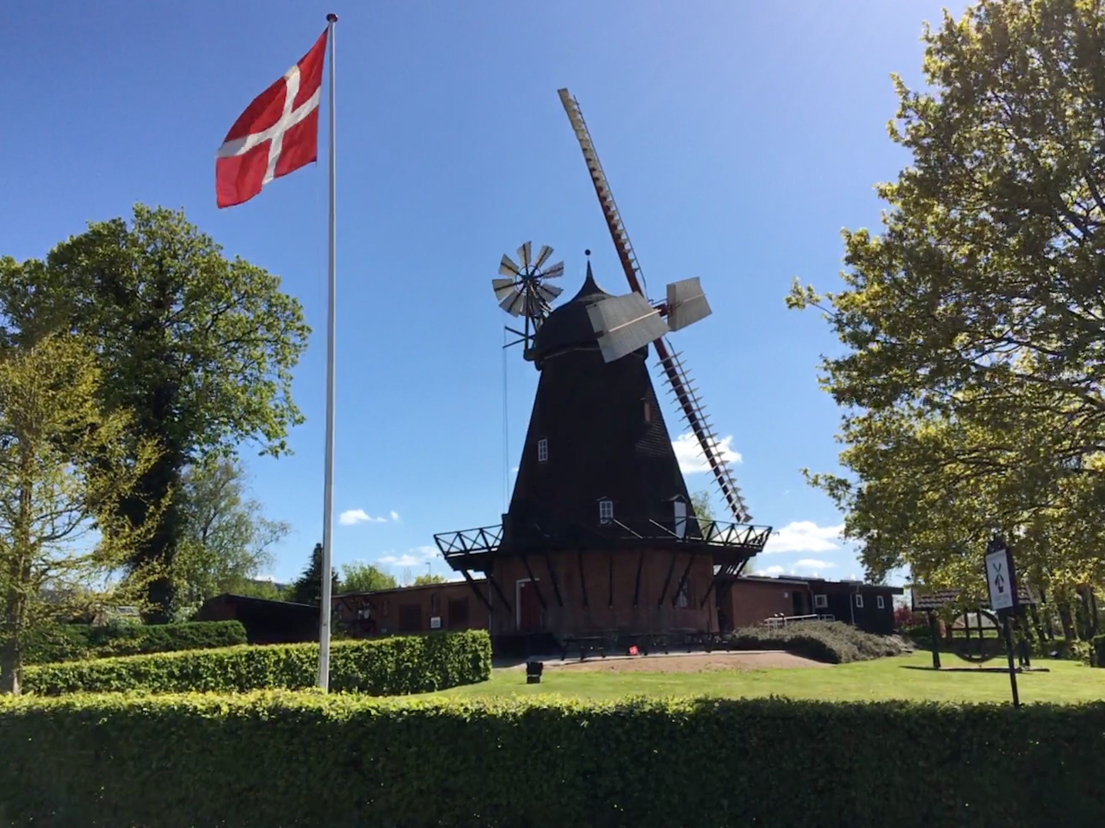 A traditional windmill with a flagpole, surrounded by well-maintained hedges and trees.